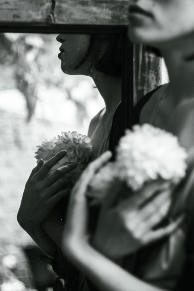Artistic black and white portrait of a woman holding flowers in front of a mirror.